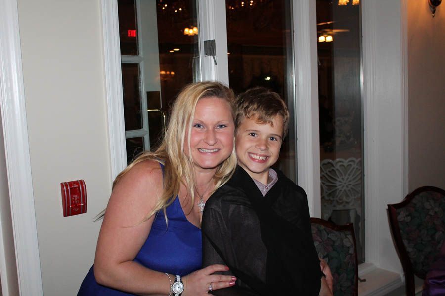 Woman in blue dress and boy in black vest smiling together indoors, near a window.