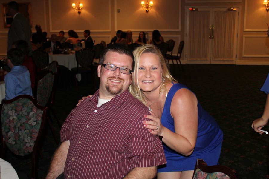Man and woman smiling, indoors. Man wearing glasses and striped shirt, woman in blue dress. Ballroom setting.