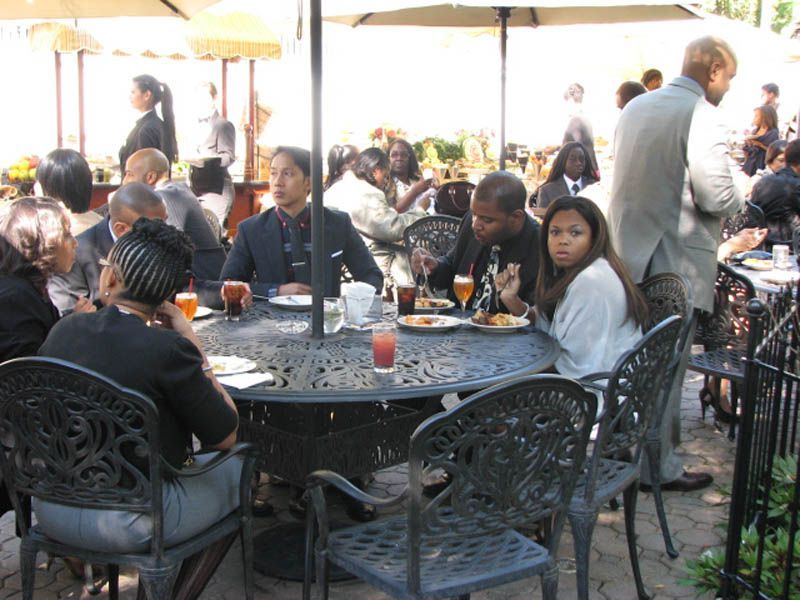 People seated around outdoor tables, eating and drinking. Sun umbrellas provide shade.