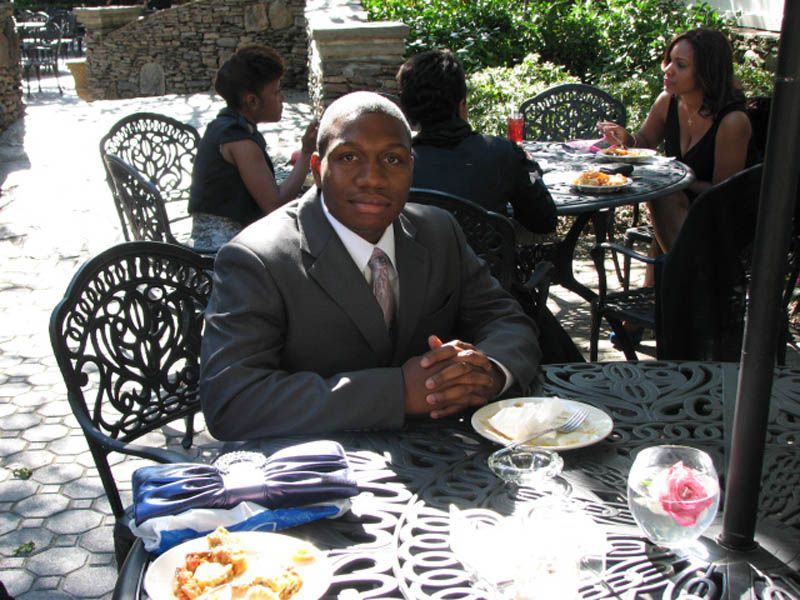 Man in suit seated at a patio table with others. Plate of food and glass of water on the table.