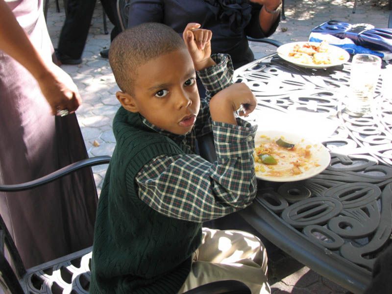 Young child at a table, wearing a green vest and plaid shirt. He is looking at the camera.