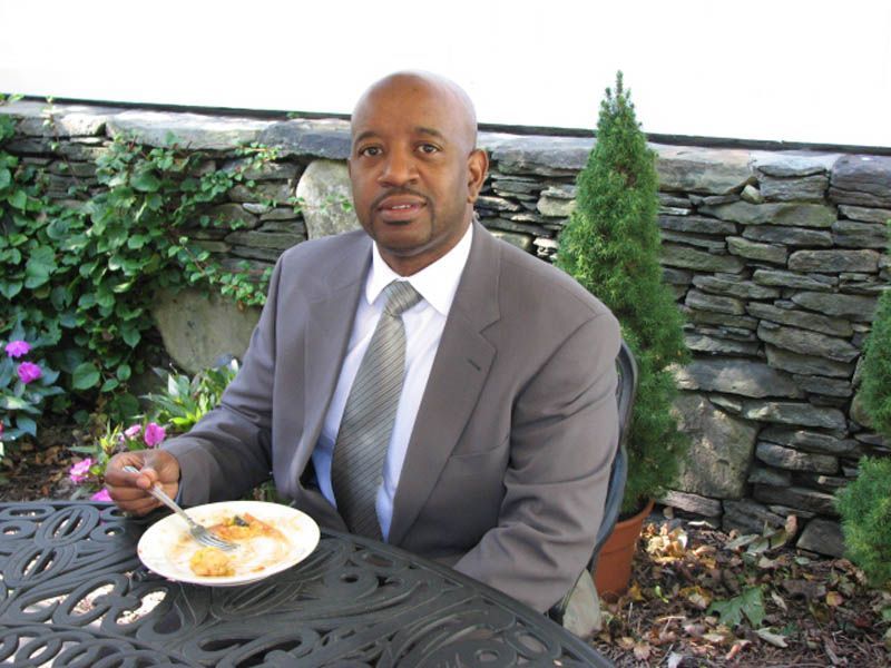 Man in suit eating at a table outdoors, stone wall and greenery in the background.