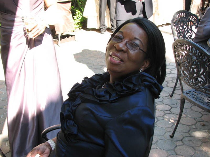 Woman smiles, wearing glasses, and a dark blue ruffled top, seated outdoors.