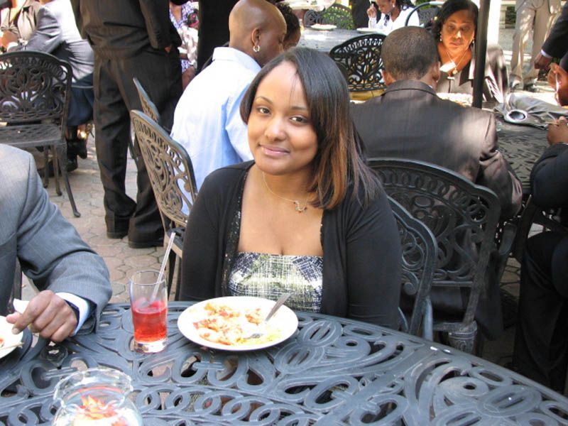 Woman at outdoor table, smiles at camera. Food and drink on table, other people in background.
