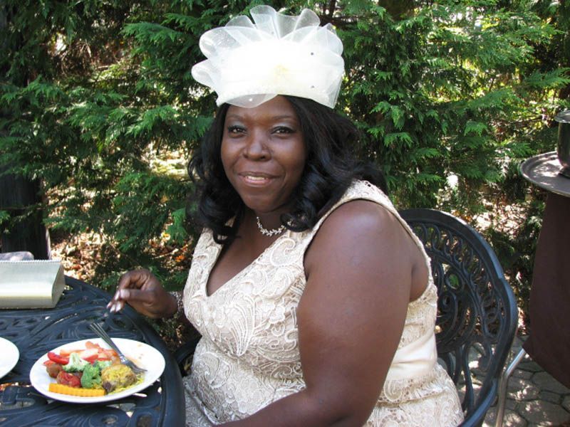 Woman wearing a cream-colored hat and dress, seated outdoors, smiling at a table with food.