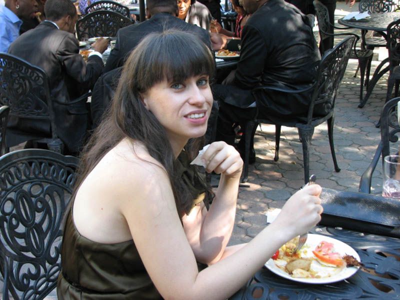 Woman eating at a patio table, brown dress, smiling, food on plate. Black metal chairs, people in background.