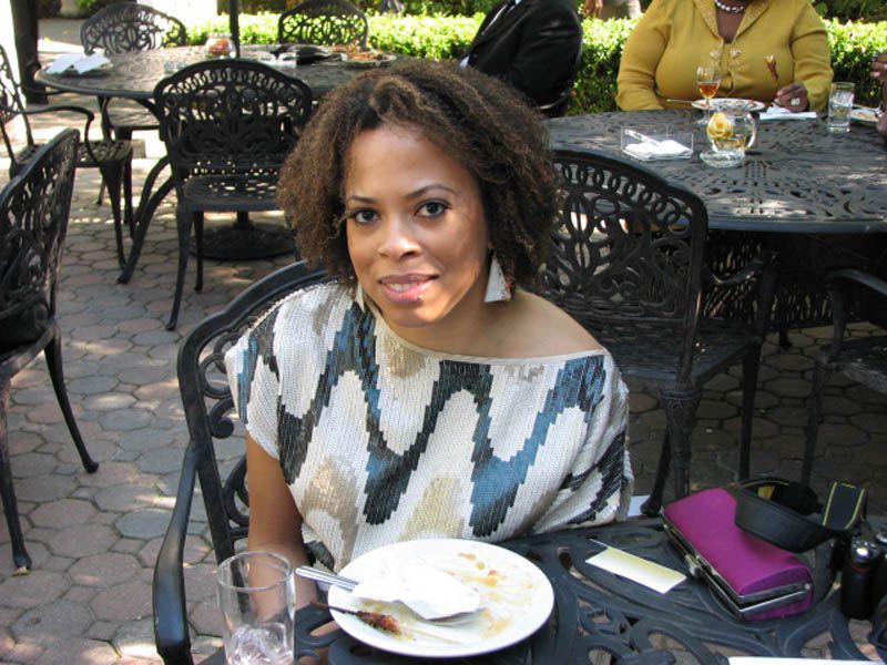 Woman seated at outdoor table, wearing patterned top, smiling. Plate and glass visible.