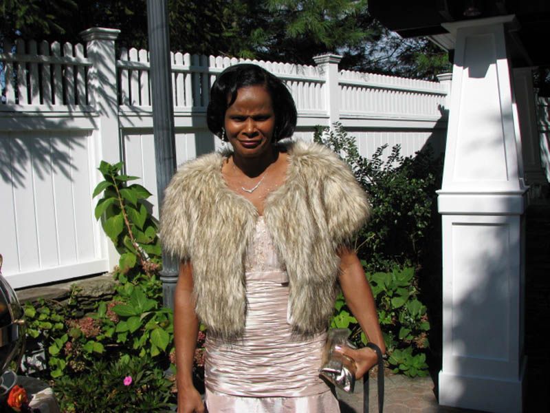 Woman wearing a fur jacket and light dress, standing outdoors in front of a white fence.