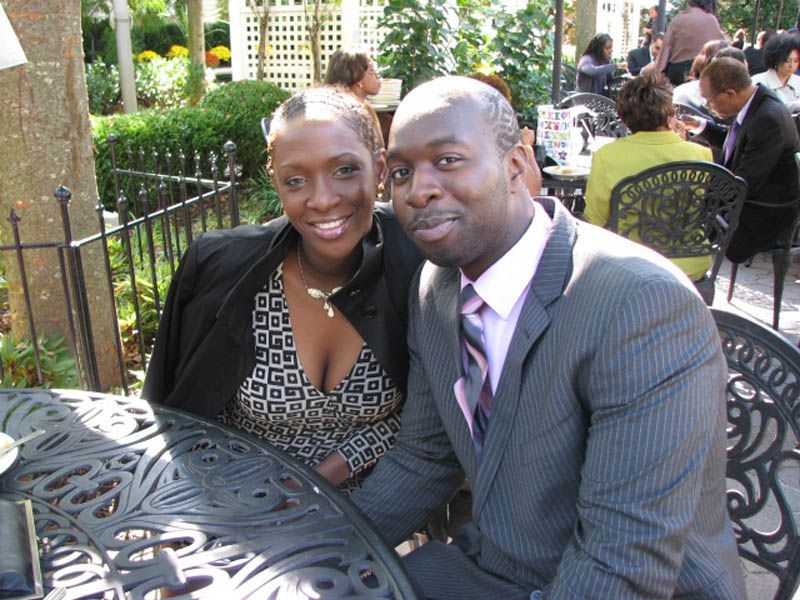 Couple smiling at a table outdoors; man in suit, woman in patterned top.