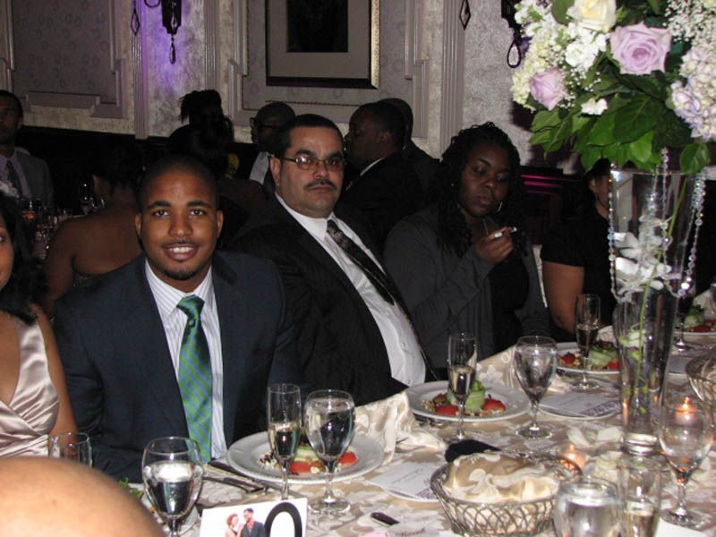 People seated at a table, one man smiling with green tie, others looking at the camera, flowers and food.