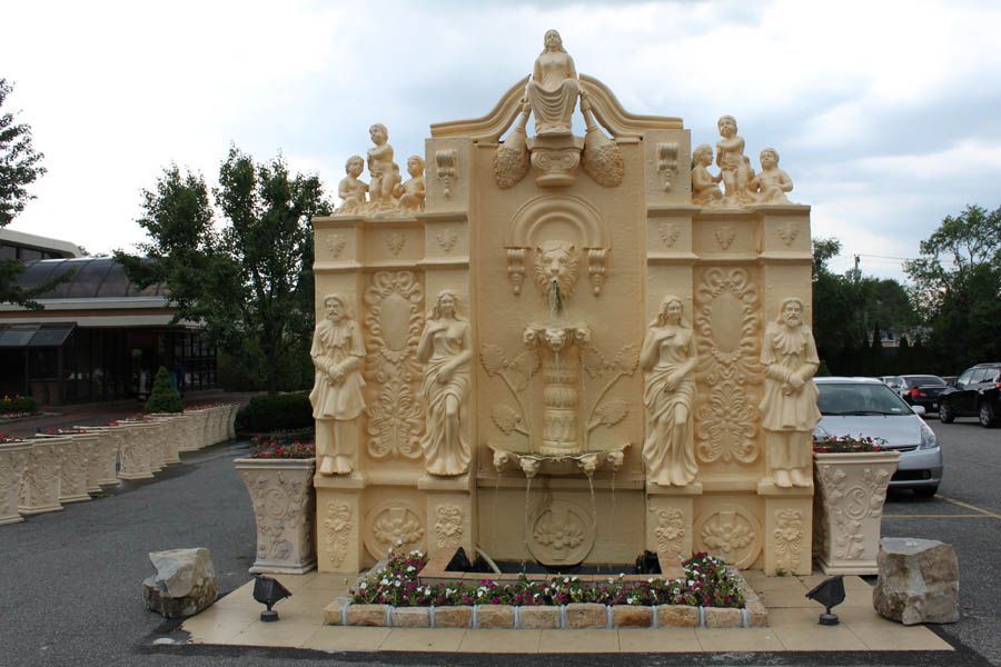 Ornate beige fountain with sculptures, surrounded by a parking lot and a building under a cloudy sky.