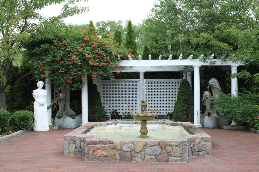 Fountain in stone basin, pergola, statues, and flowering tree in a garden setting.