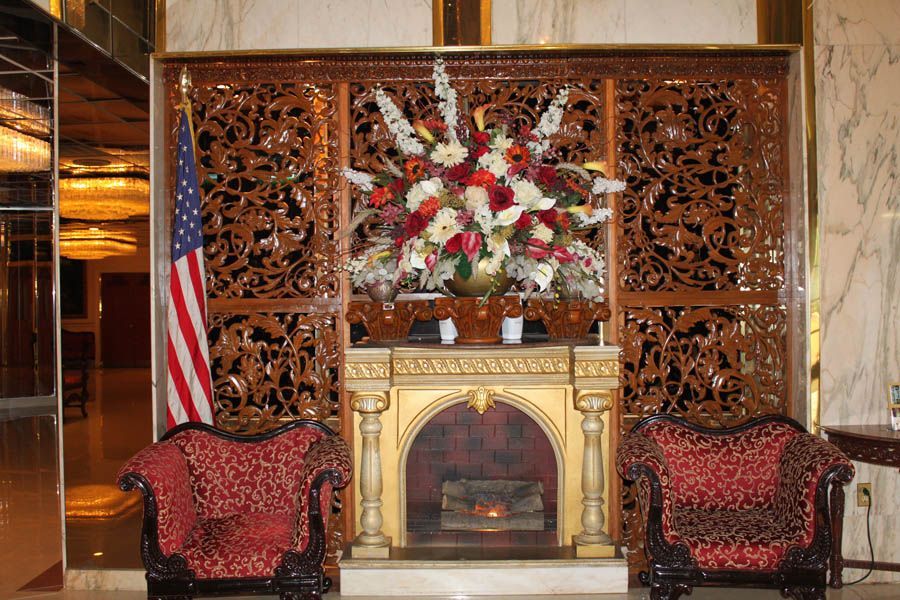Elegant lobby with carved wood panels, fireplace, flower arrangement, US flag, and ornate chairs.
