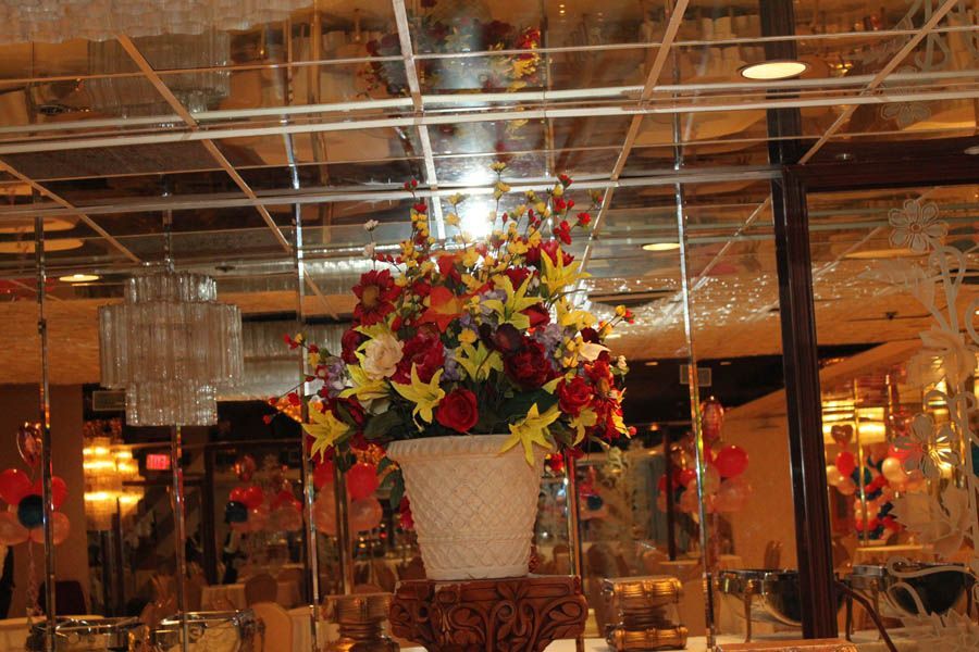 Floral arrangement in a white vase on a carved pedestal in a mirrored banquet hall, with a chandelier in the background.