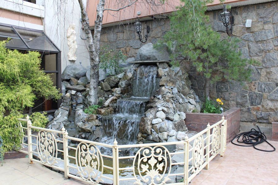 Small waterfall feature behind a decorative fence, made of rocks, with a stone wall backdrop.