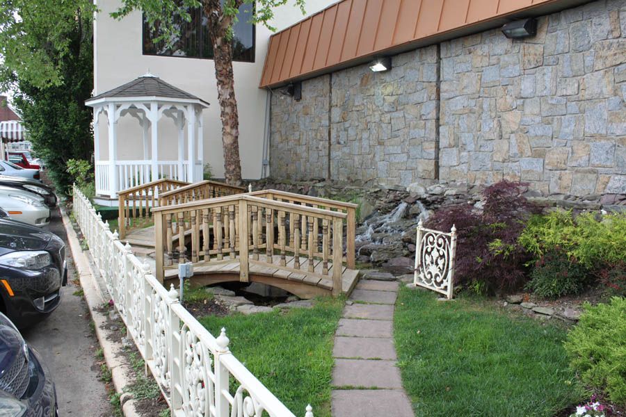 Wooden bridges and gazebo in a garden next to a stone building and a white fence, cars parked nearby.
