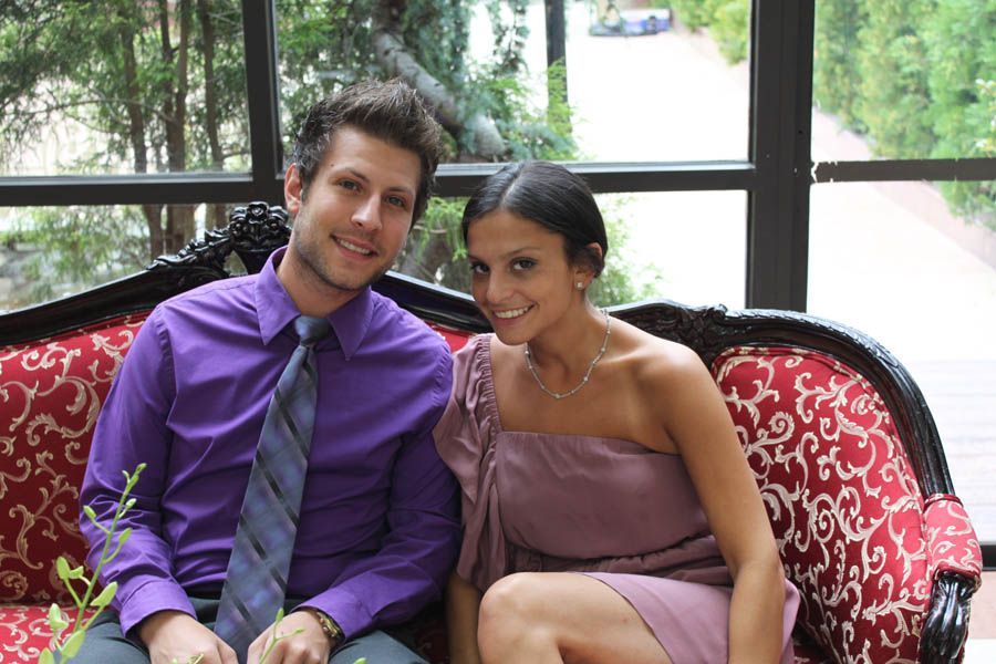 Couple smiling on a red patterned couch near a window. Man in purple shirt, woman in lavender dress.