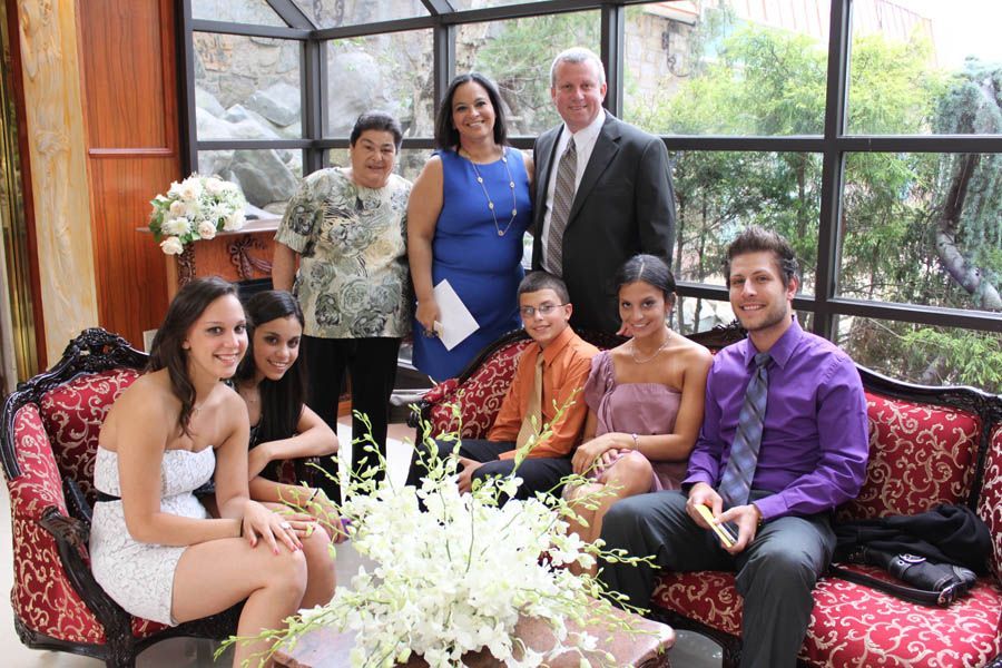 Family group poses near a floral arrangement and ornate sofa. Some smile, near windows with greenery.