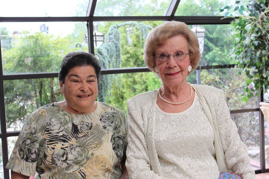 Two women smiling, posing inside by a large window. One in white jacket, pearl necklace, the other in a patterned blouse.