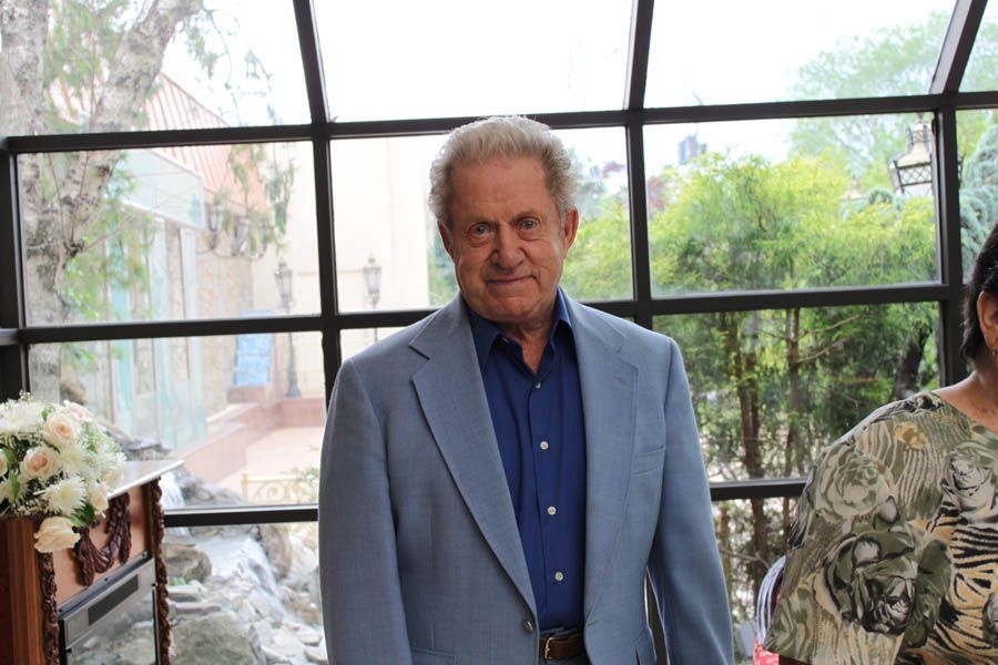 Man in blue suit, smiling slightly, standing near a window with a garden view.