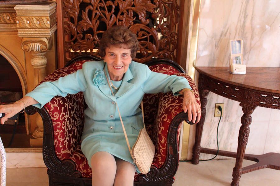 Woman in blue suit sits in ornate chair; detailed wooden paneling and table in background.