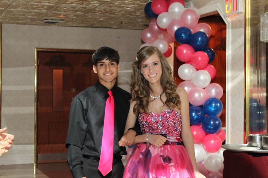 Couple at a formal event; boy in black shirt and pink tie, girl in pink sequin dress; balloons in background.