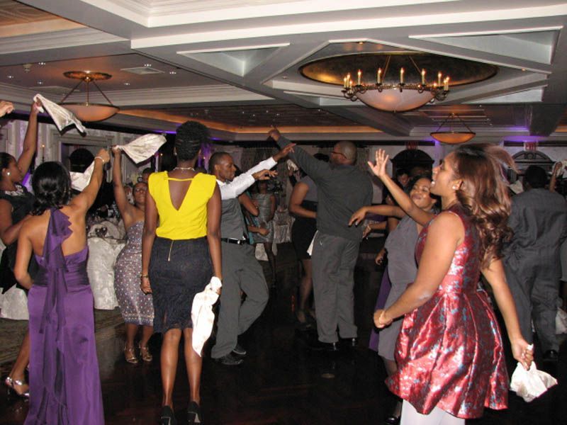 People dancing enthusiastically in a ballroom with a chandelier; some are holding napkins, and many are smiling.