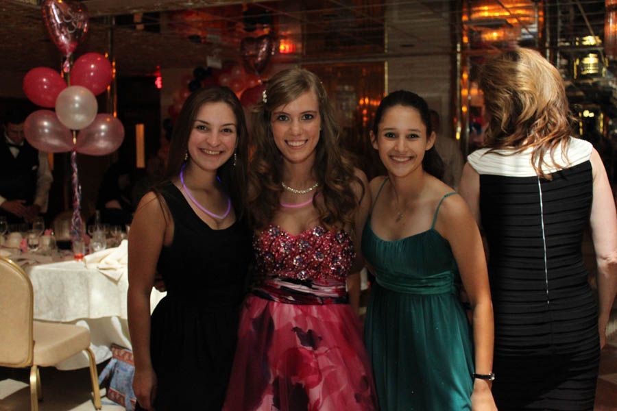 Three young women in formal dresses pose at a celebration, pink and clear balloons in background.