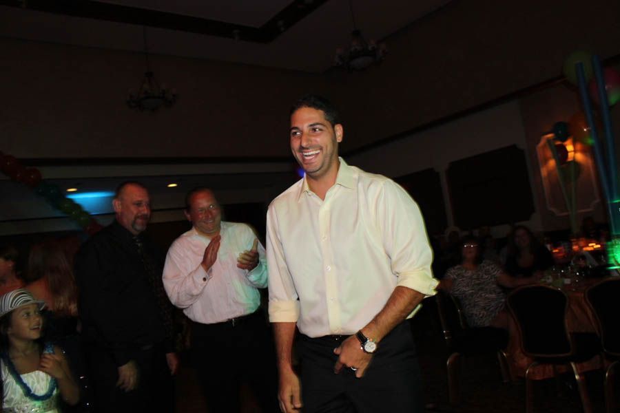 Man in cream shirt smiles, dancing at an event, with two men clapping behind him. Dimly lit room.
