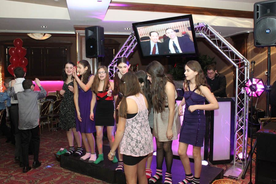 Teenagers singing and dancing on a stage in a party setting, with a DJ and a video screen playing behind them.