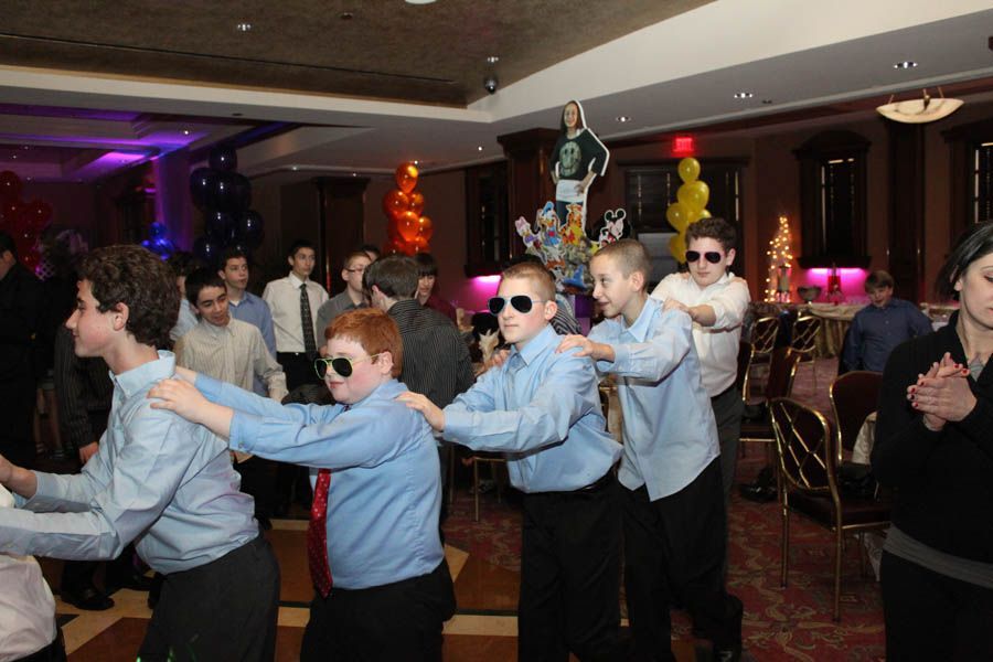 Boys in sunglasses, blue shirts, and black pants dancing in a room with balloons and decorations.