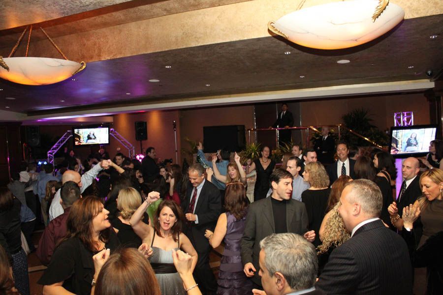People dancing at an event in a dimly lit room with televisions on the wall and decorative lights.