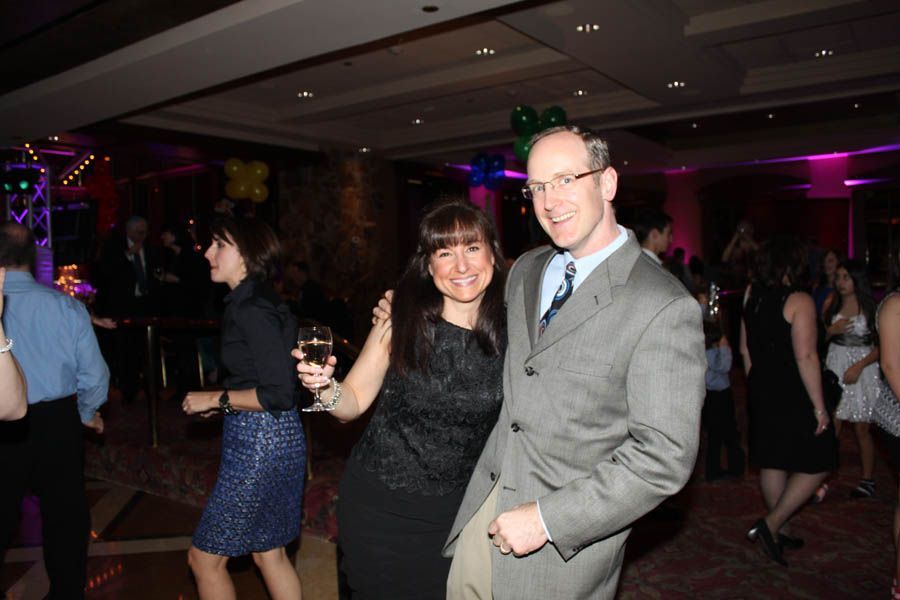People dancing in a brightly lit ballroom. A woman smiles with a drink, and a man in a suit poses beside her.
