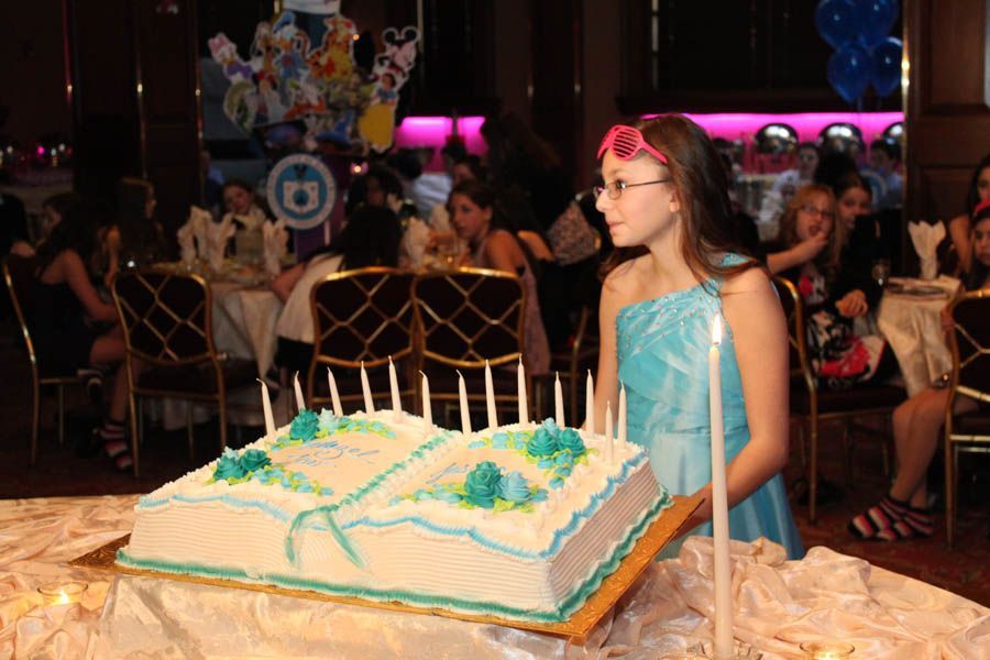 Girl in blue dress with a birthday cake shaped like a book, lit candles. Party setting.