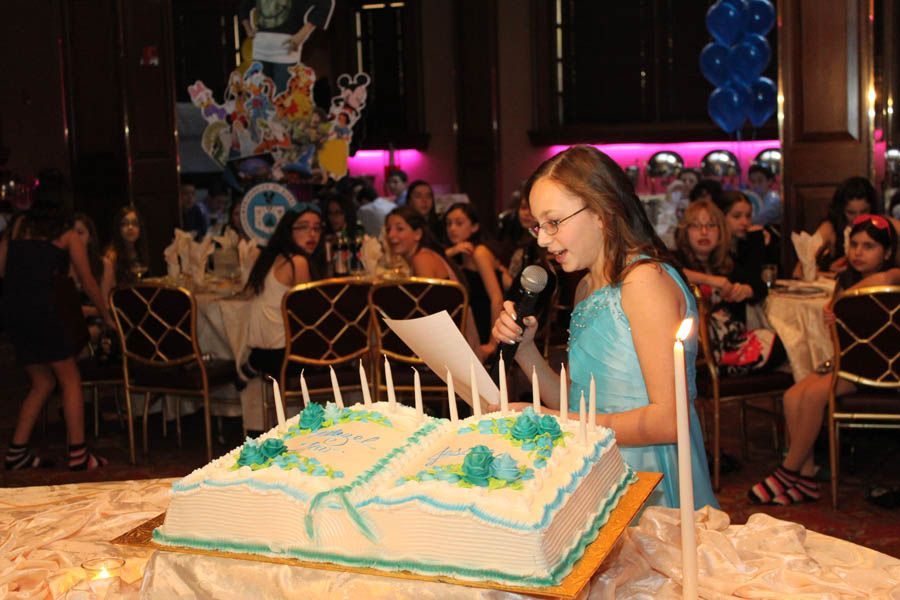Girl with microphone reading by a cake shaped like a book, lit candles. Party guests in background.