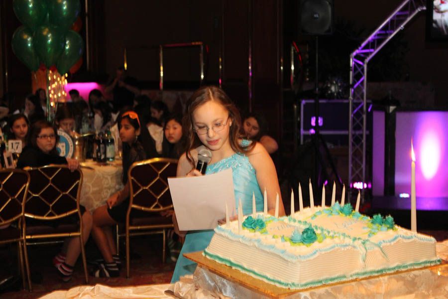 Girl giving speech at party; cake with candles in foreground; guests seated at tables.