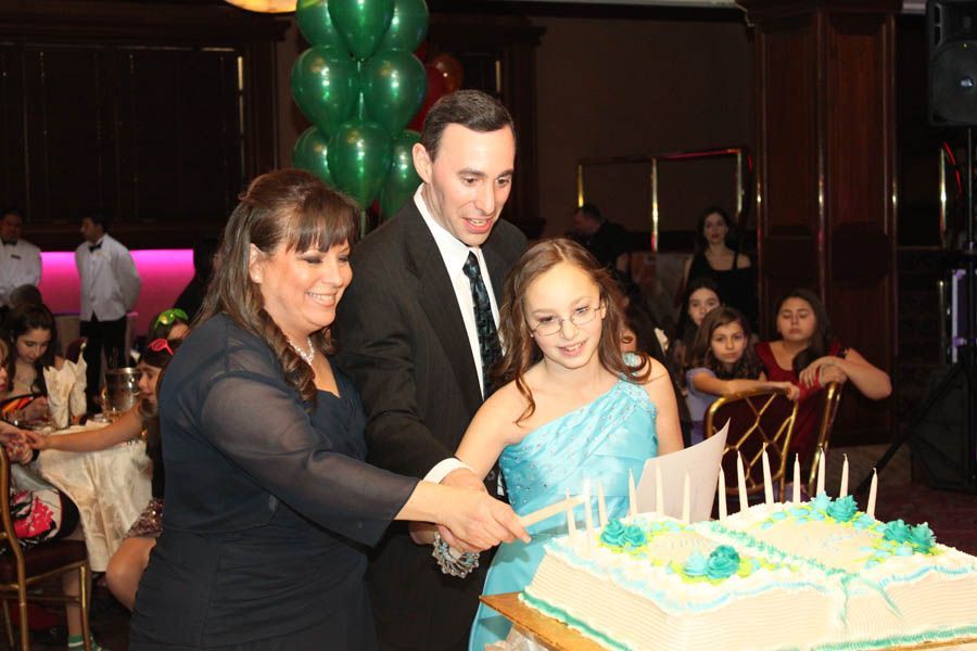 Family cuts a teal frosted birthday cake with lit candles at a party with green balloons.