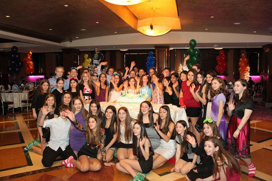 Large group of young people celebrating around a birthday cake in a ballroom, smiling and posing.