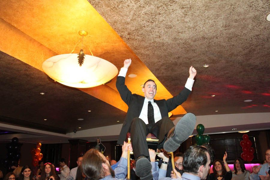 Man in suit is lifted up by a crowd, arms raised. Party setting, happy expression.
