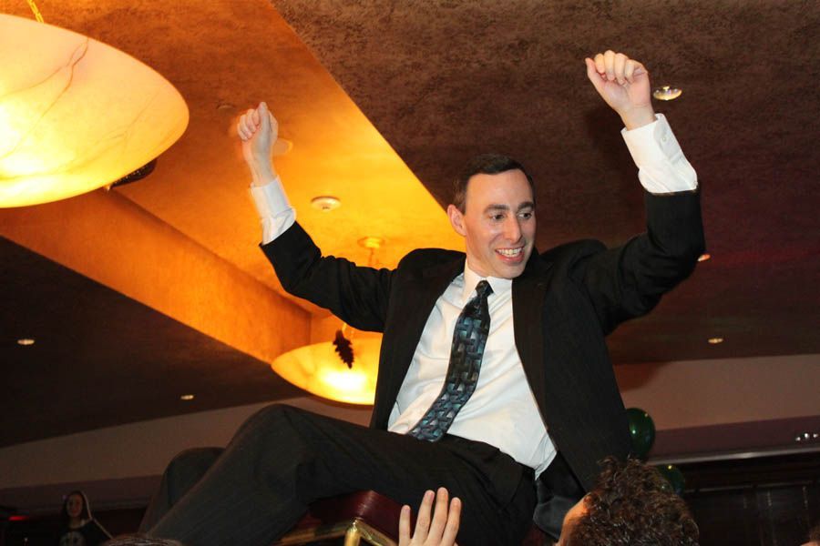 Man in a suit raised on shoulders, arms up, celebrating indoors.