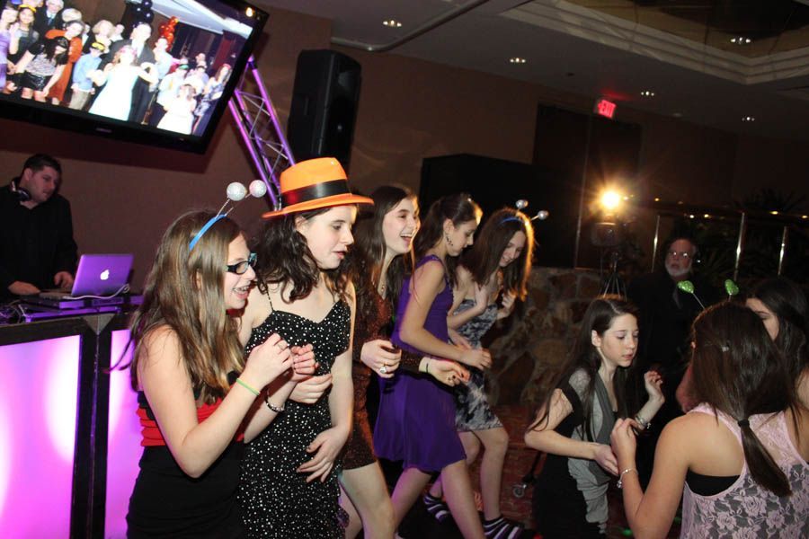 Group of girls dancing at a party, illuminated by pink and yellow lights, a DJ in the background.