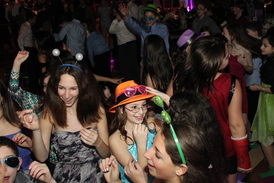 People dancing at a party, wearing hats and glasses. Dimly lit with some smiling faces and raised arms.