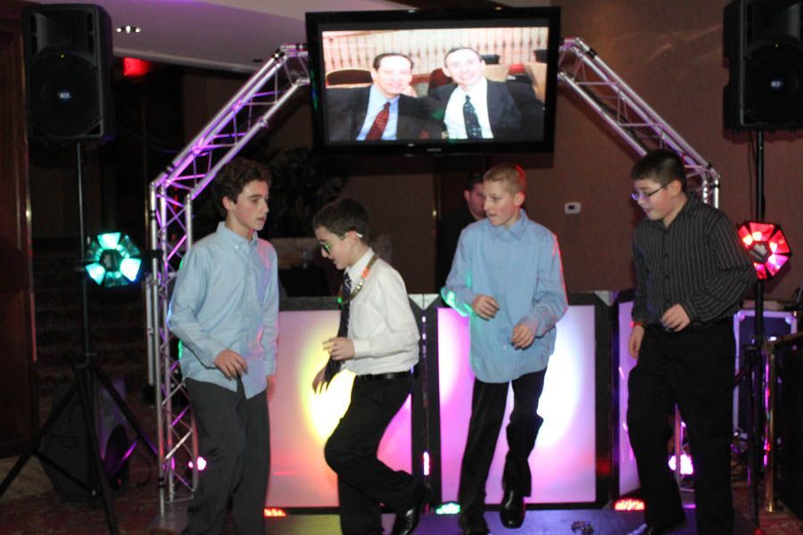 Four boys dancing in front of a DJ booth with a screen displaying two men, speakers, and colored lights.