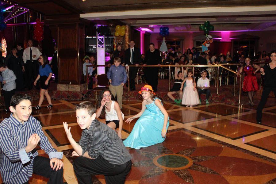 People dancing and posing on a dance floor at a party. Some are children. Balloons and a fancy setting are visible.