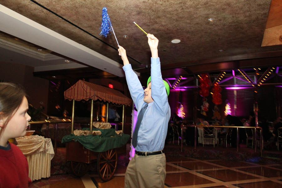 Man in blue shirt and tie, holding up a wand and a blue flag, entertaining guests at a party.