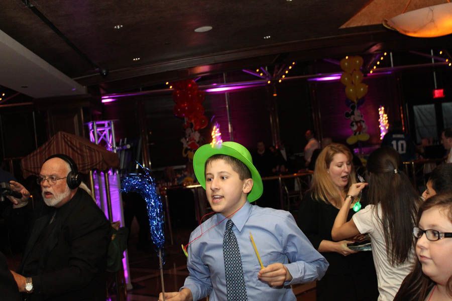 Teen boy dancing with party hat at an event, surrounded by others, DJ in headphones.