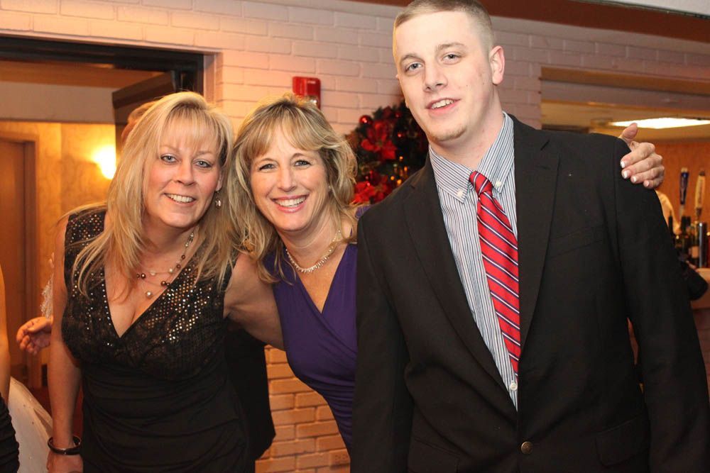 Three people smiling, posed close together indoors; one in a black dress, one in a purple dress, and one in a suit.