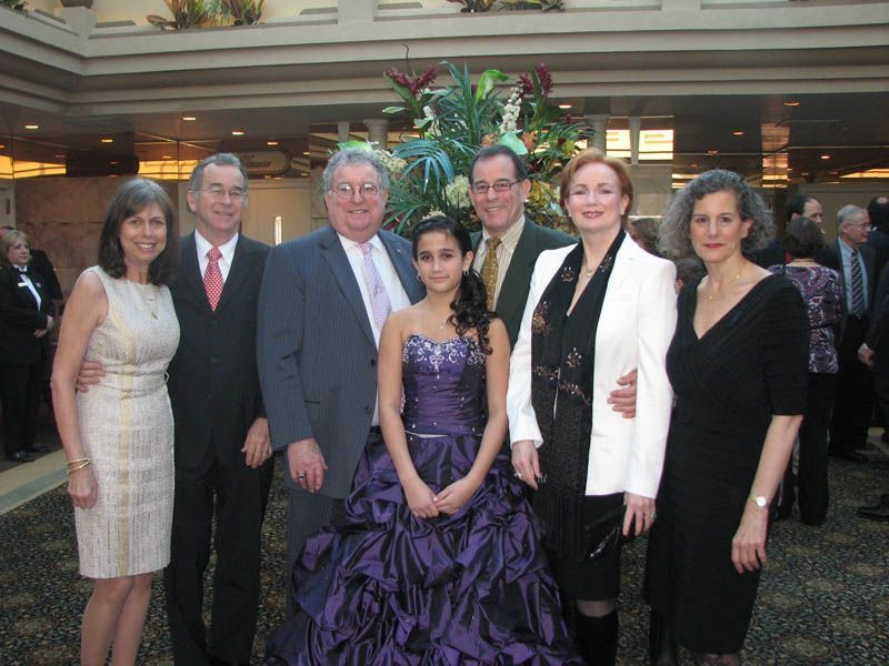 Group poses in formal attire; woman in purple ball gown is central. Indoor setting, various skin tones.