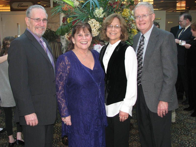 Four people smiling, posing in front of a floral display, at an event.