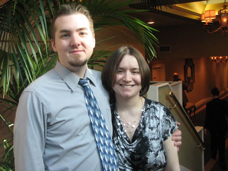 Man with tie and woman in formal wear, arm-in-arm, smiling, in a well-lit hall with greenery.